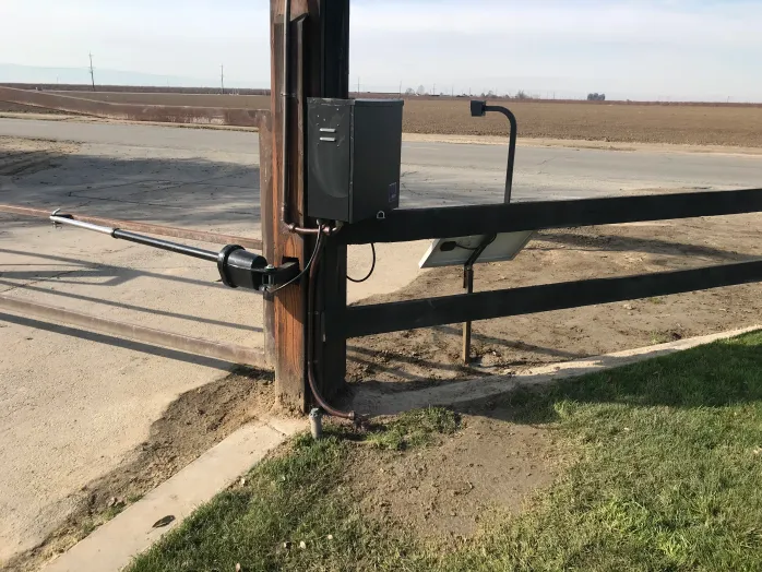 Black metal farm gate with electric opener beside a dirt road in a flat rural field